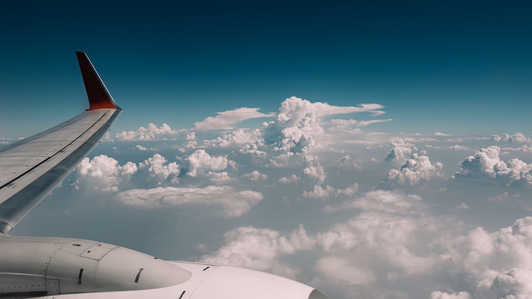 An airplane window view at cruising altitude showing clouds and wing, with a smart ring on a finger in the foreground